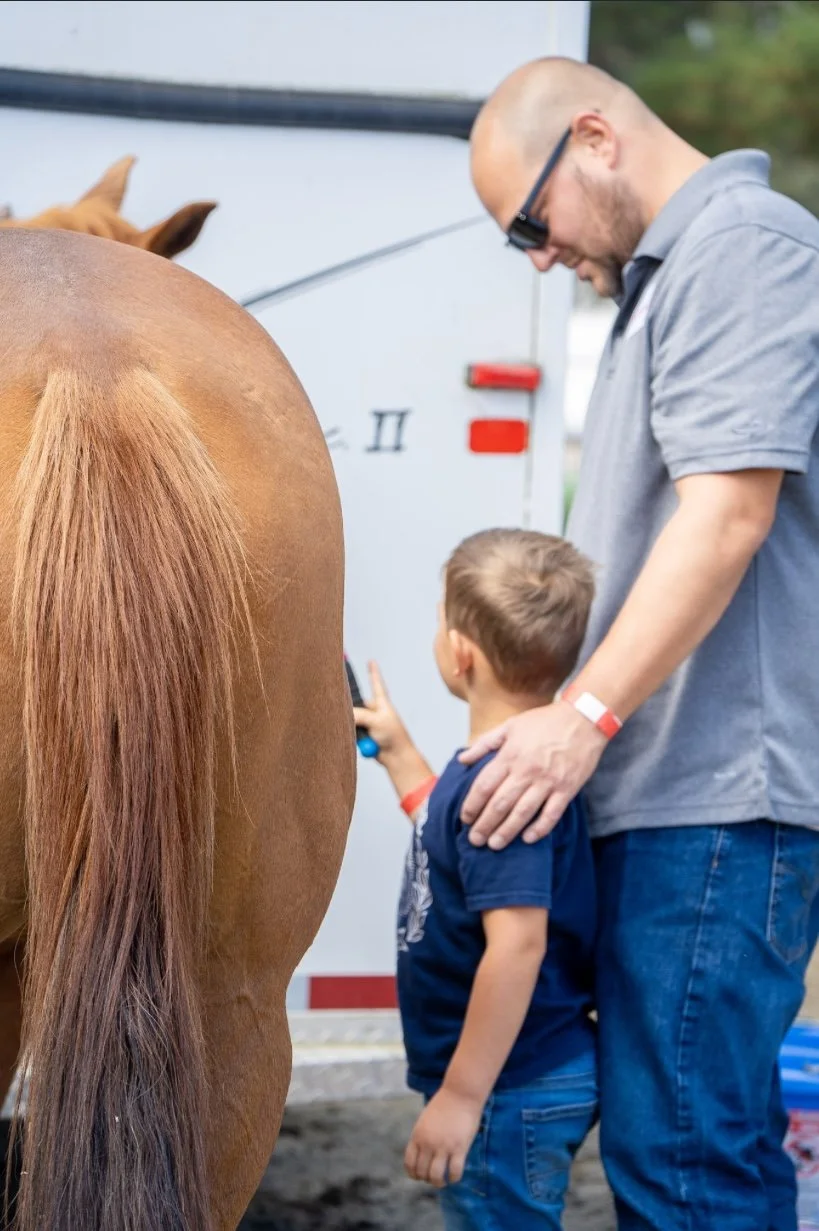 A man and a young boy interact with a brown horse, with the man's hand resting on the boy's shoulder, at an outdoor event or fair.