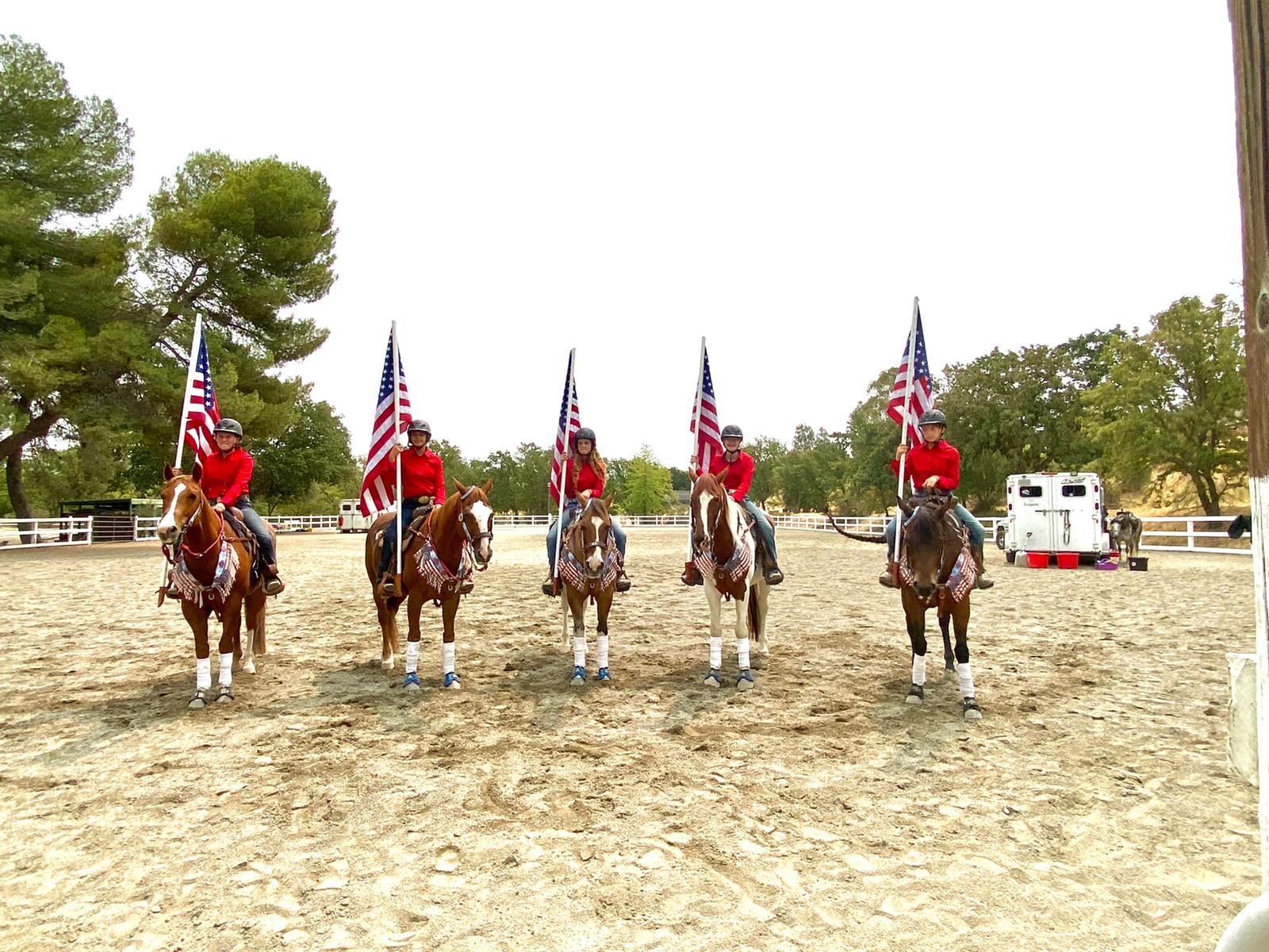 Five people riding horses, each holding a large American flag, wearing red shirts and riding gear, in an outdoor riding arena with trees and a white fence in the background.