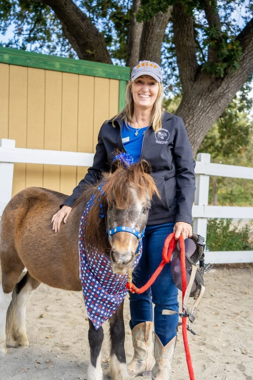 A woman standing outdoors next to a small horse, with her hand on the horse's back. The woman is smiling, wearing a gray cap and a black jacket with a logo. The horse is decorated with a patriotic bandana and a matching headband, and is attached to a