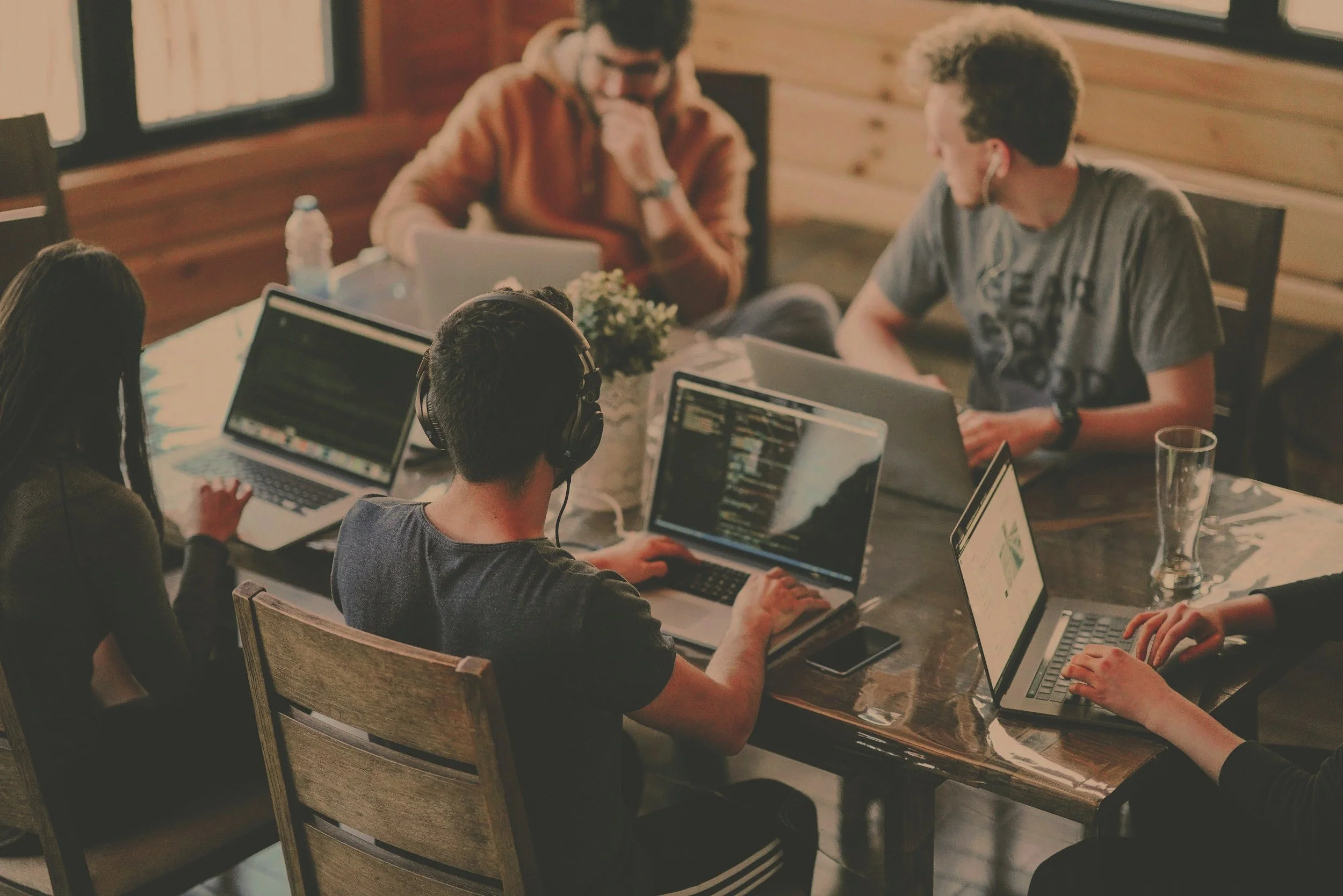 A group of five young people working together at a wooden table with laptops and headphones in a cozy, wood-paneled room.