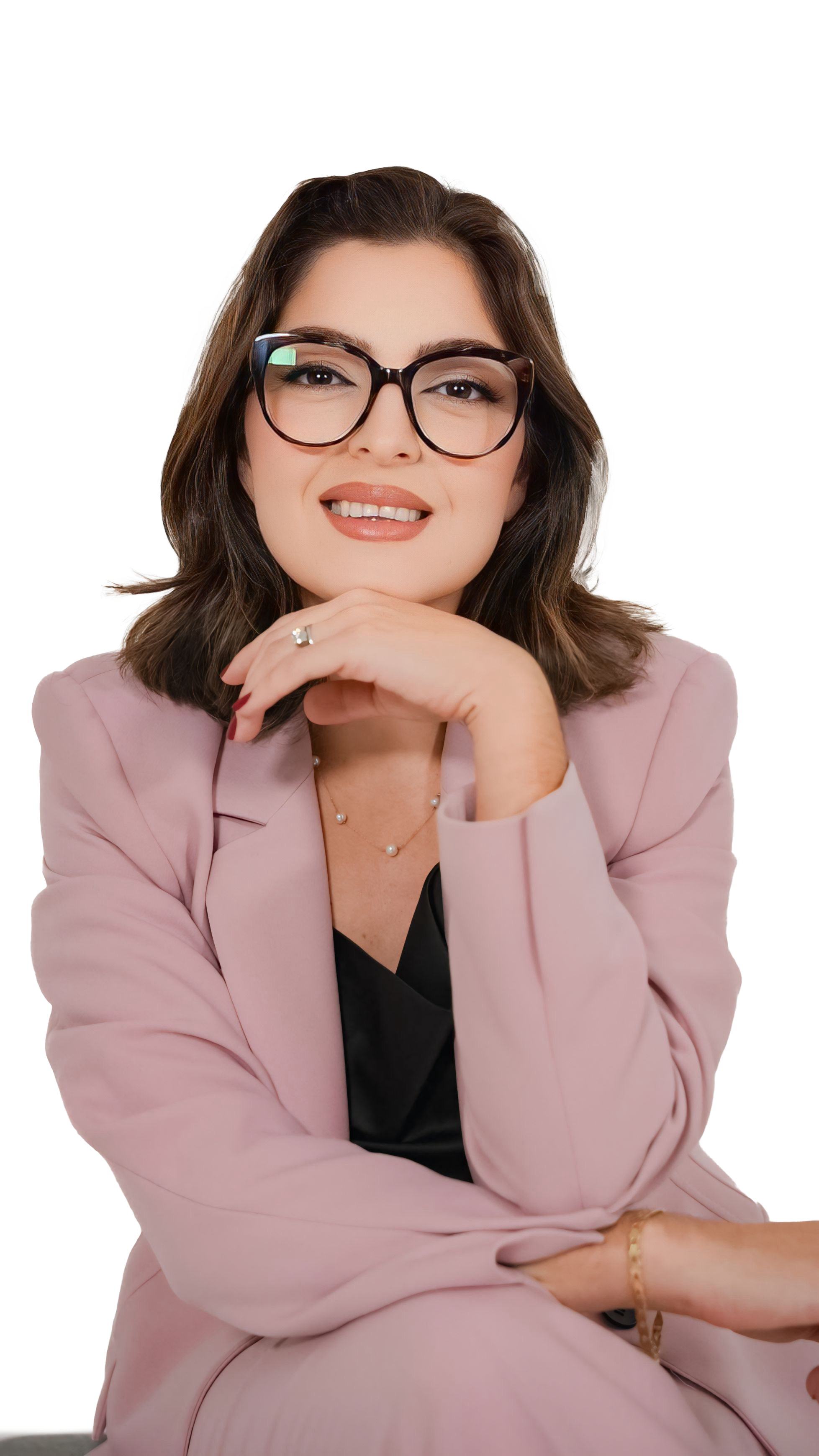 A woman with shoulder-length brown hair wearing glasses, a pink blazer, a black top, and jewelry. She is sitting with her chin resting on her hand, smiling at the camera.