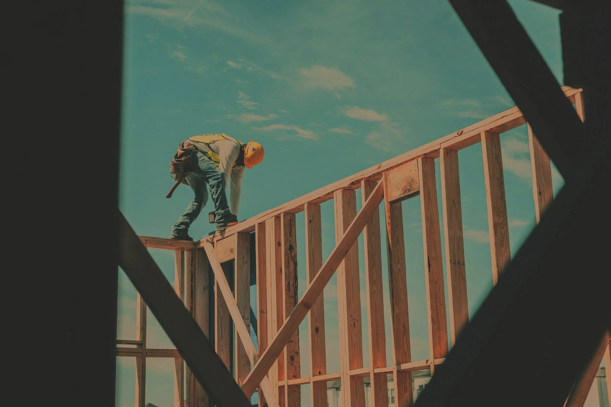 A construction worker wearing a yellow safety vest, orange hard hat, and jeans is working on the wooden frame of a building under a partly cloudy sky.