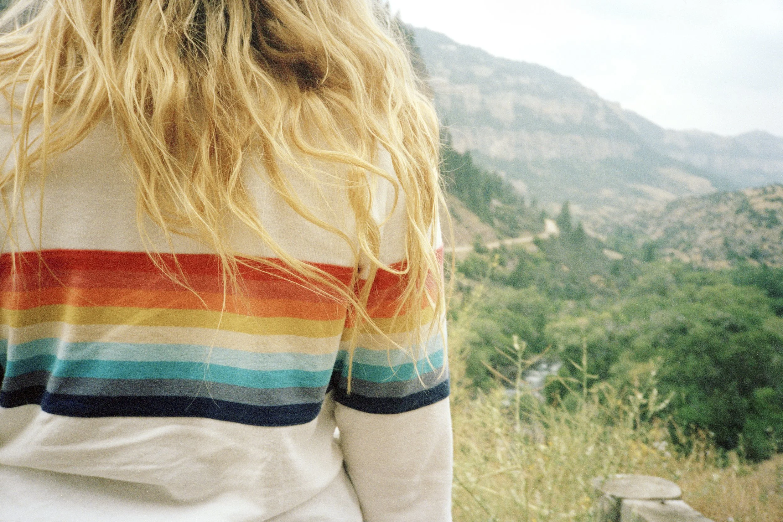 Close-up of a person's torso wearing a white shirt with colorful horizontal stripes in red, orange, yellow, green, blue, and black, standing outdoors with a mountainous landscape in the background.