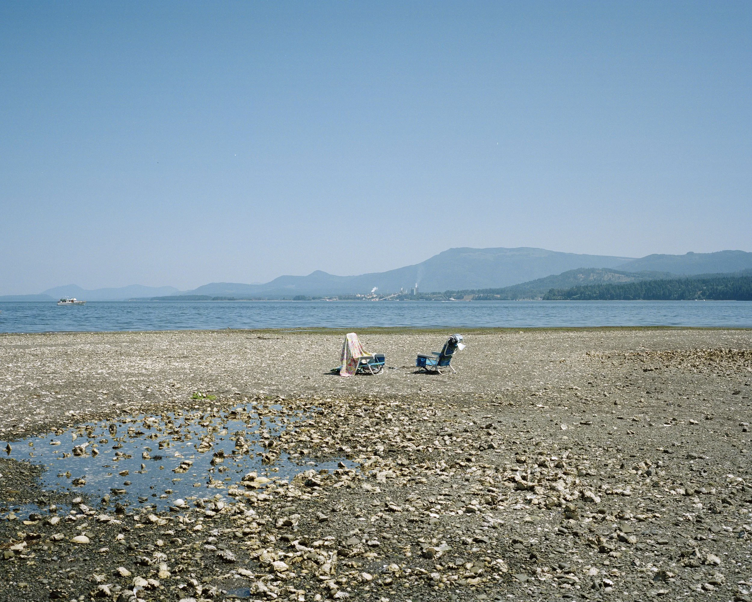 A lakeshore with two chairs and a towel, boats on the water, distant mountains, and a clear blue sky.