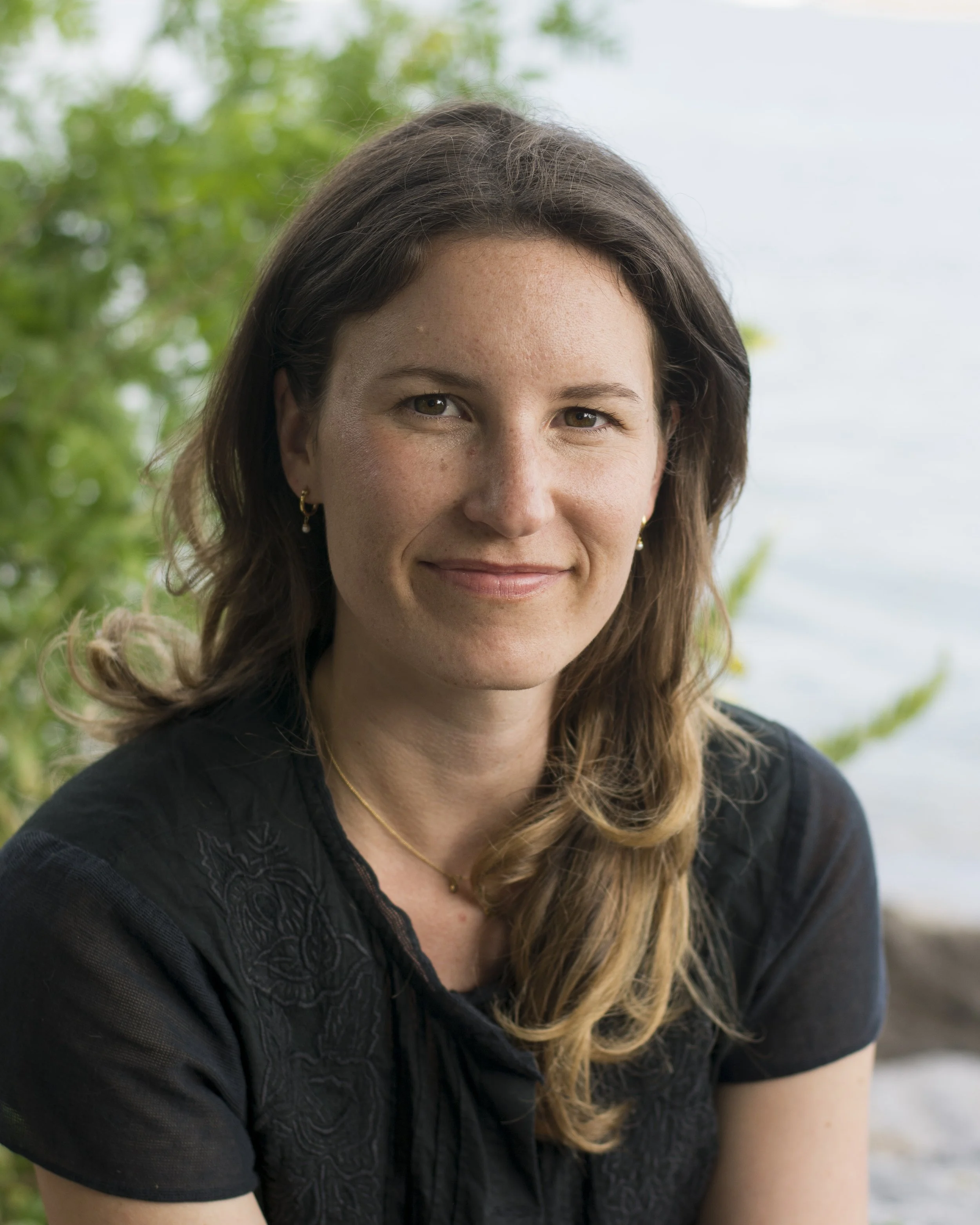 Close-up of a woman with brown hair, wearing a black top and earrings, outdoors with green foliage and a cloudy sky in the background.