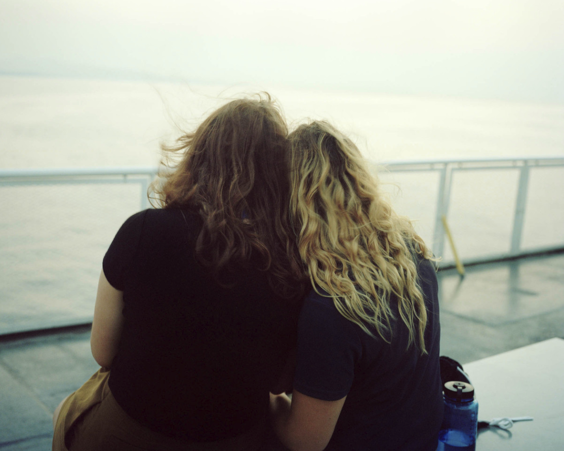 Two women with curly hair sitting close together by the water, viewed from behind, on a boat deck during the day.