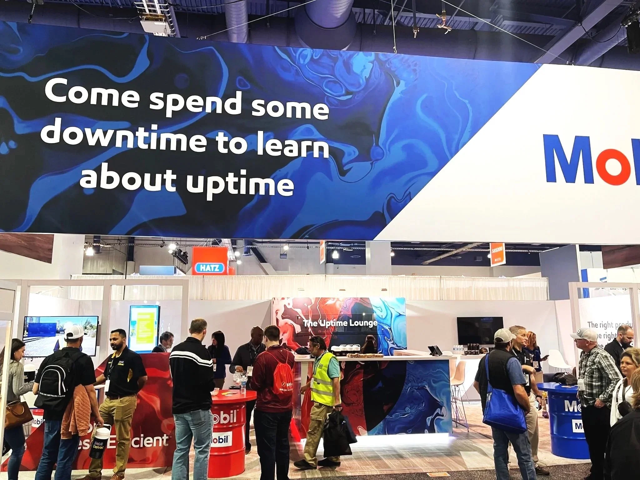 A busy trade show with a large overhead sign that says, 'Come spend some downtime to learn about uptime,' above booths and people mingling. The booths include Mobil and other automotive-focused brands, with displays and informational counters.