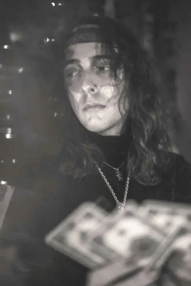 Black and white photo of a person with long, wavy hair, wearing a dark top and jewelry, holding cash in front of them, with a serious expression.