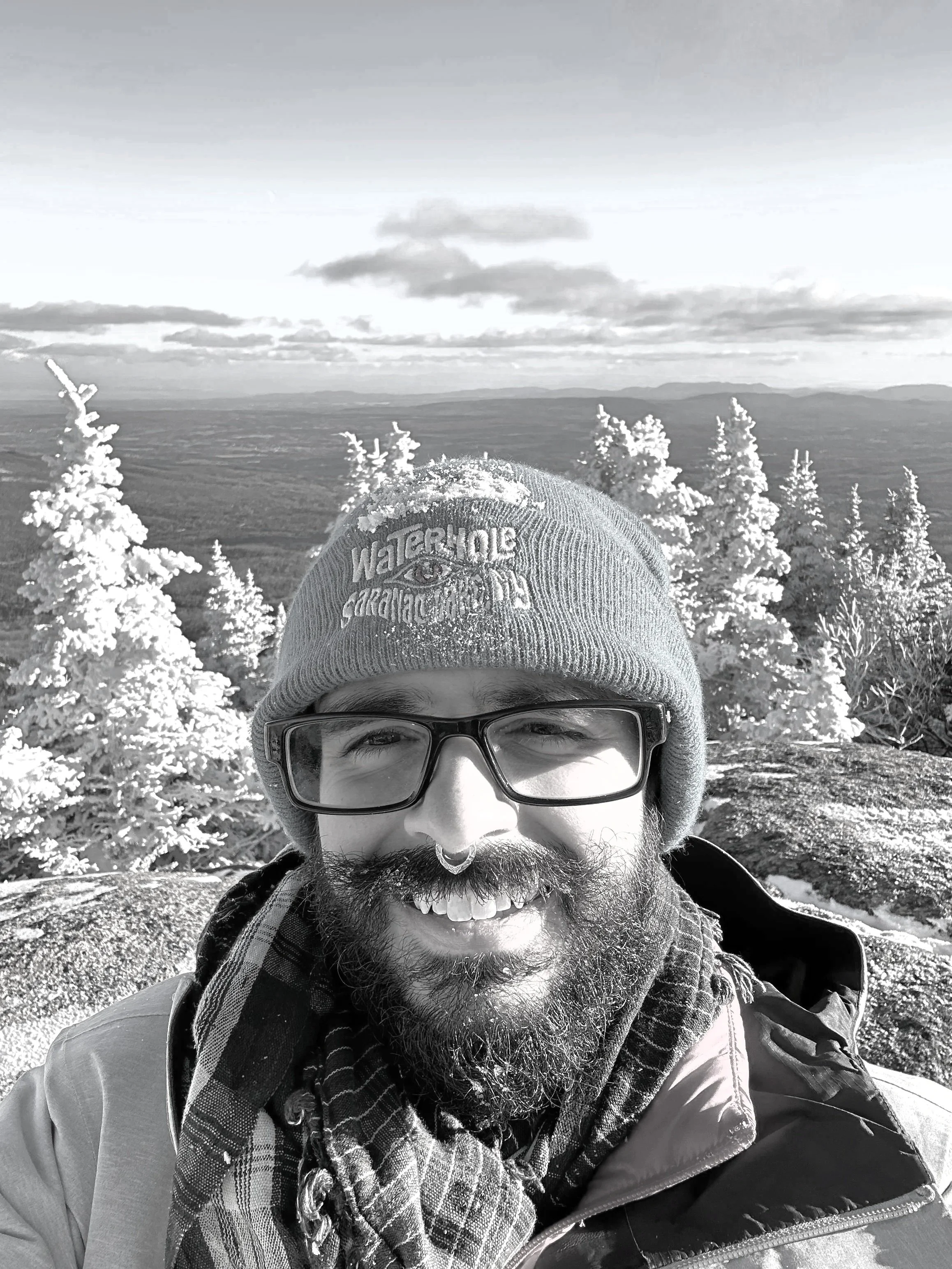 A person with a beard, wearing glasses, a water-resistant hat, and a jacket, stands outdoors in a snowy landscape with snow-covered trees and mountains in the background. The person is smiling and appears to be enjoying a winter hike or outdoor adventure.