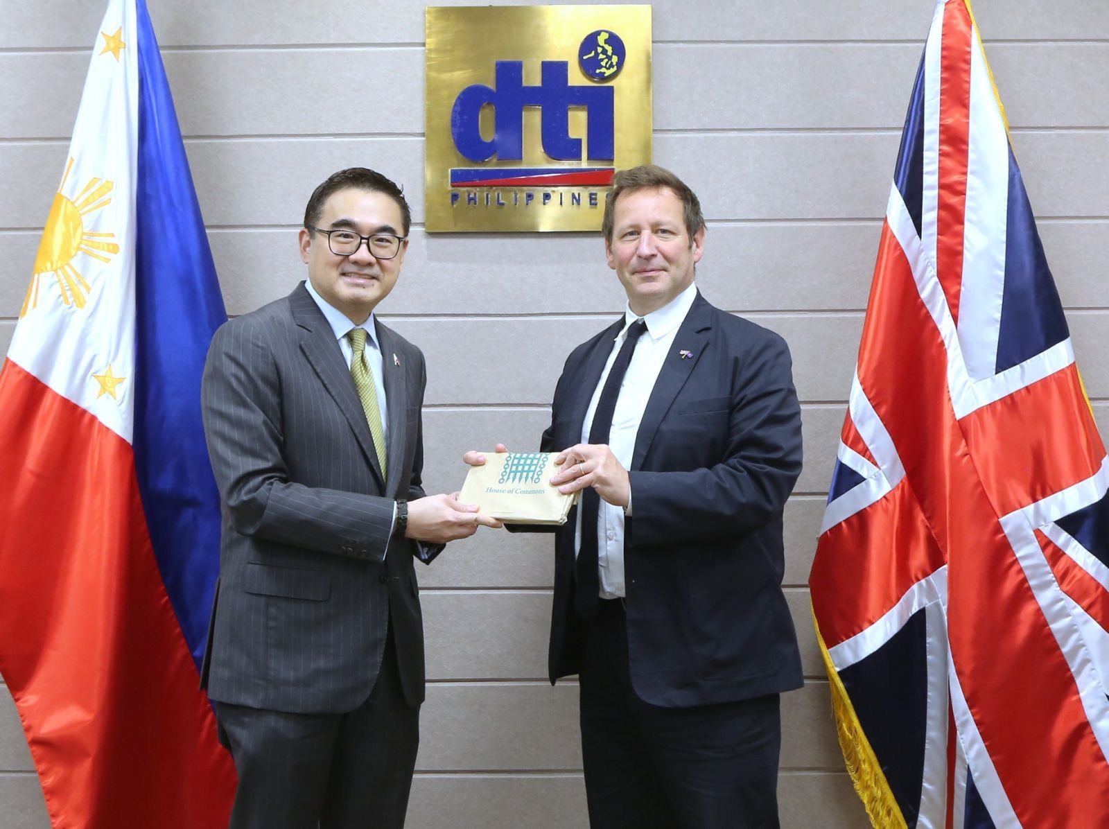 Two men in suits standing in front of flags and a sign that reads 'dtI PHILIPPINES'. The man on the left is holding a ballot band while the man on the right is receiving it. The Filipino flag is on the left and the British flag is on the right.
