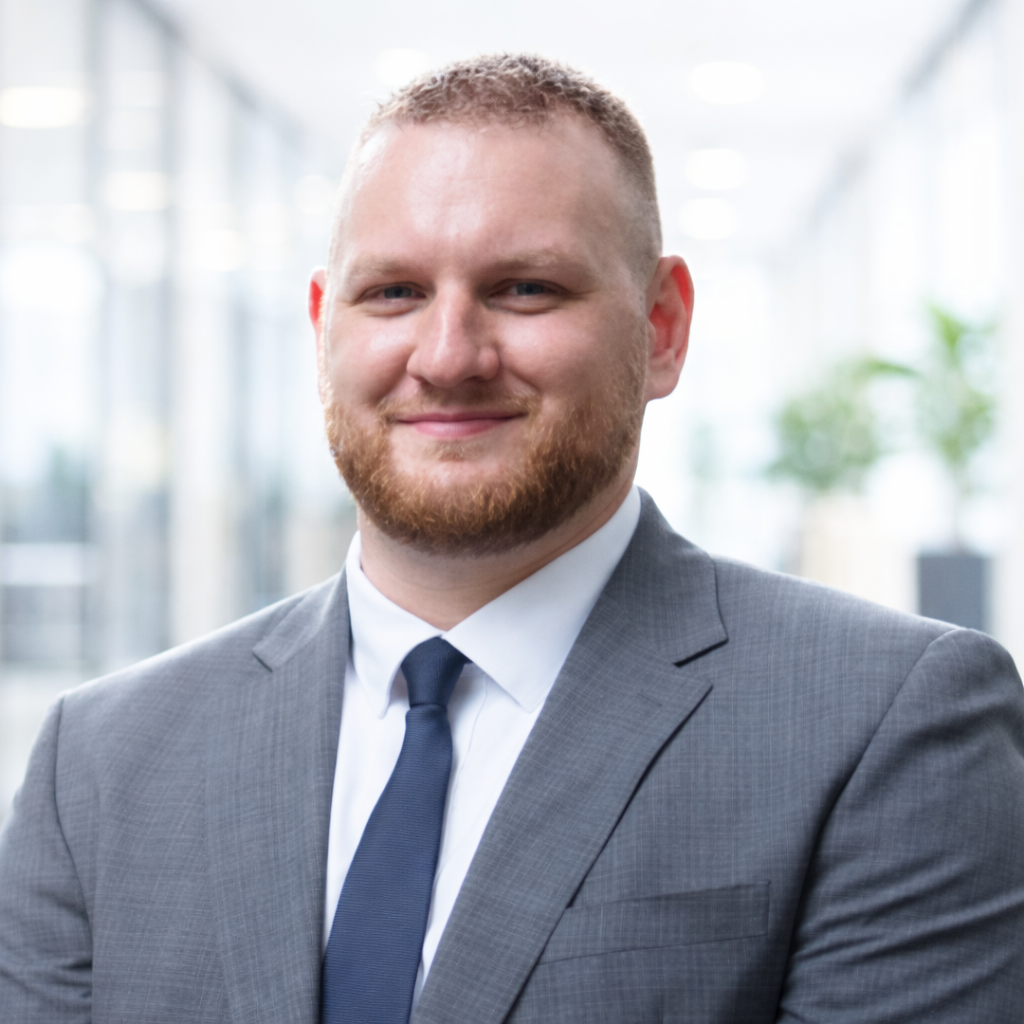 Headshot of a man with short red hair, blue eyes, and a beard, wearing a dark gray blazer and a black shirt against a plain white background.