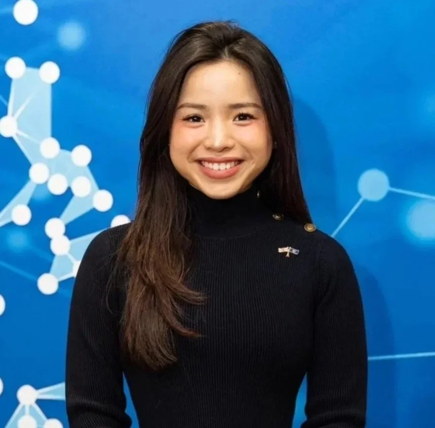 A young woman with long dark hair smiling at the camera, wearing a navy blue sweater over a white collared shirt, standing in front of a blue background with a network graph design.