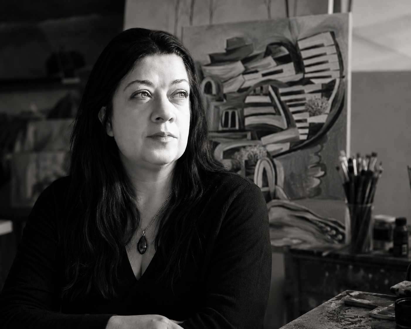 A woman with long dark hair looking thoughtfully to her left, sitting in an artist's studio with paintings and art supplies in the background.