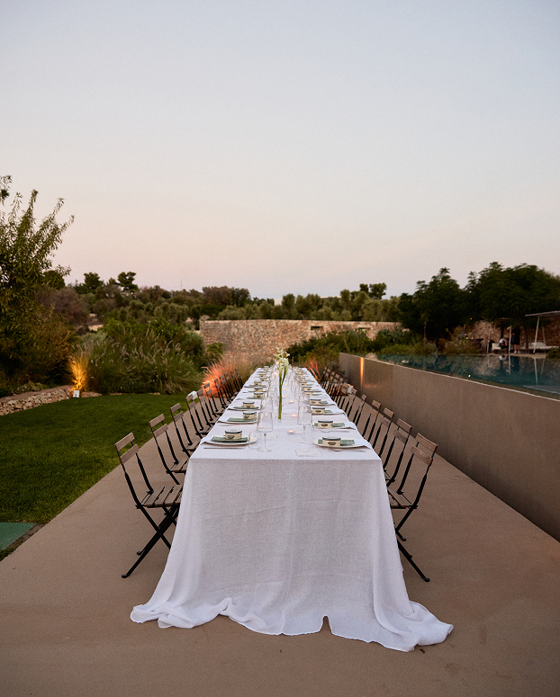 Long outdoor dining table with white tablecloth, set with plates, glasses, and napkins, surrounded by black chairs, overlooking a landscaped garden and sunset sky.