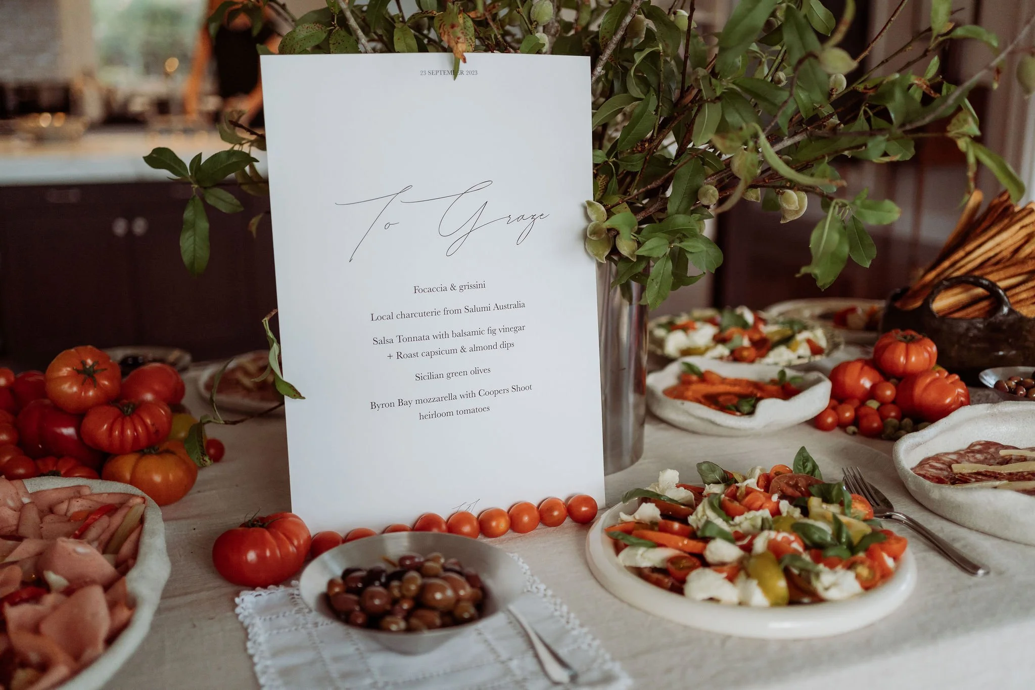 A table set with fresh tomatoes, salads, and various appetizers, with a white menu card in a metal vase and some greenery.