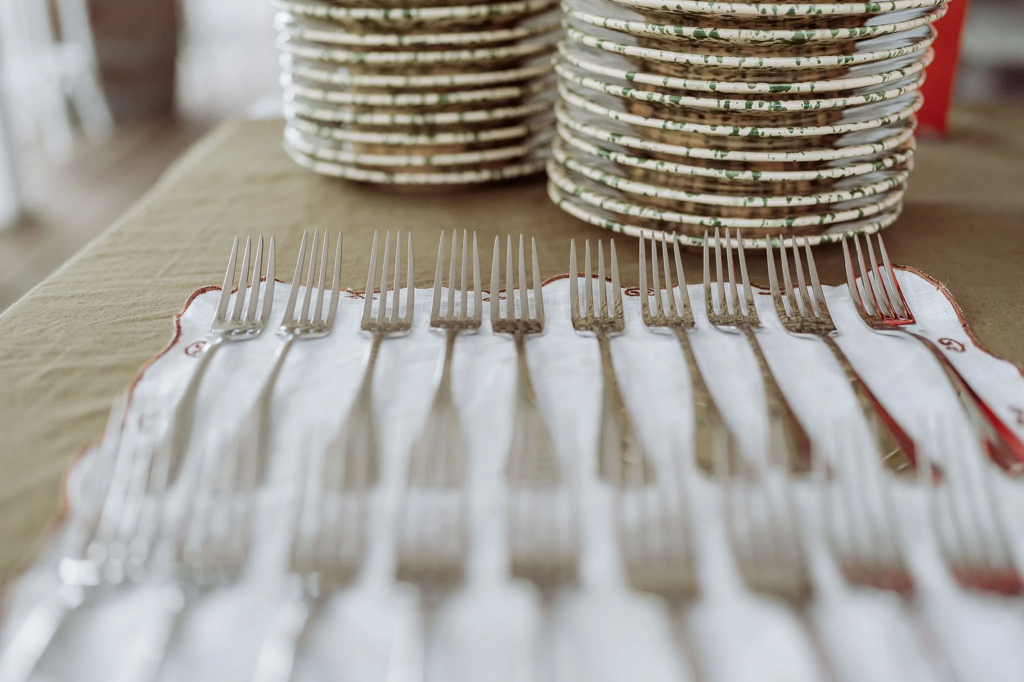 Multiple stainless steel forks arranged on a white cloth napkin with red embroidery, in front of stacked paper plates on a table.