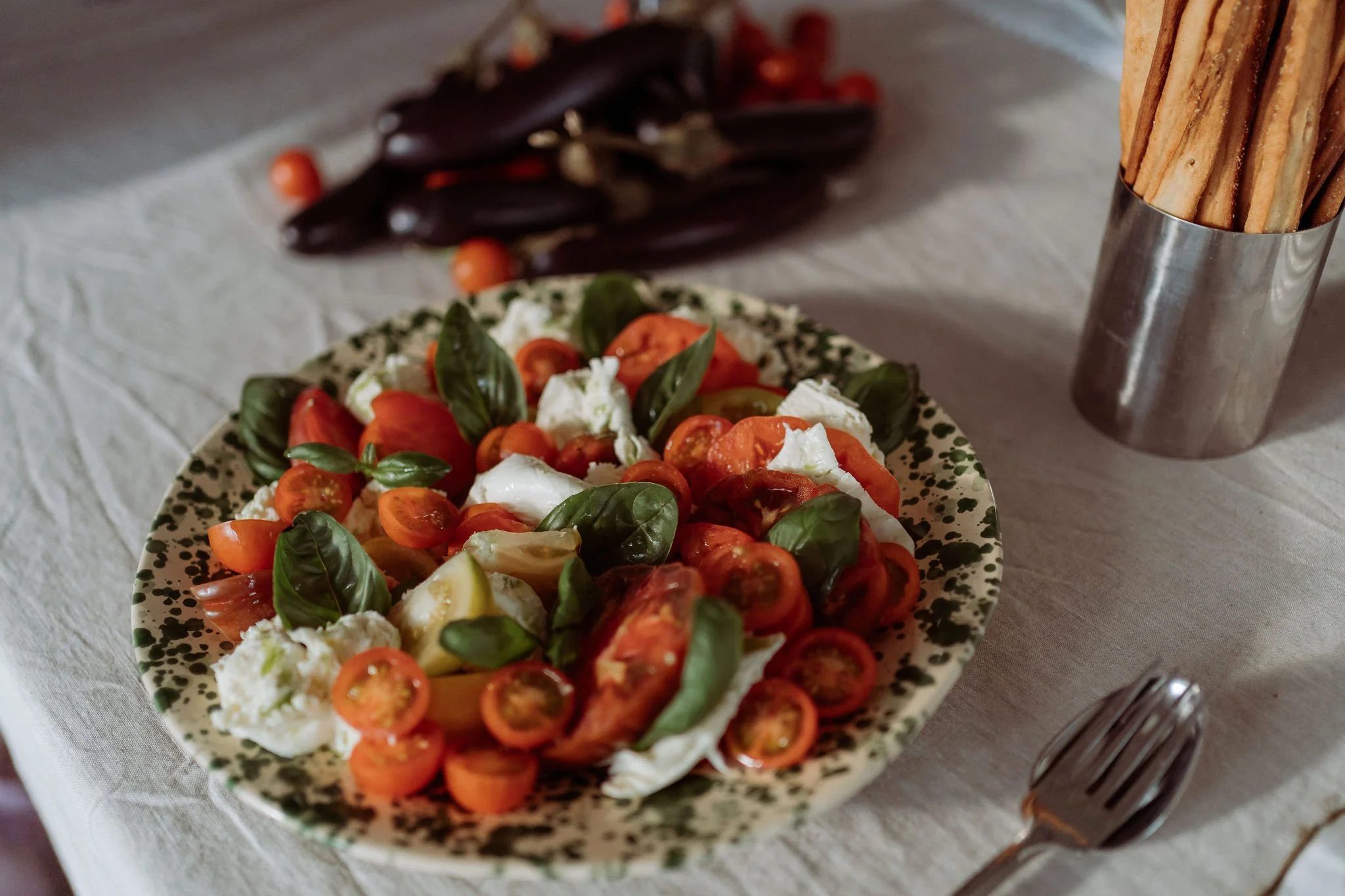 A plate of fresh Caprese salad with cherry tomatoes, basil leaves, and mozzarella cheese on a patterned plate. In the background, there are eggplants with cherry tomatoes on a cloth-covered table, and on the right, a metal container holding breadstic