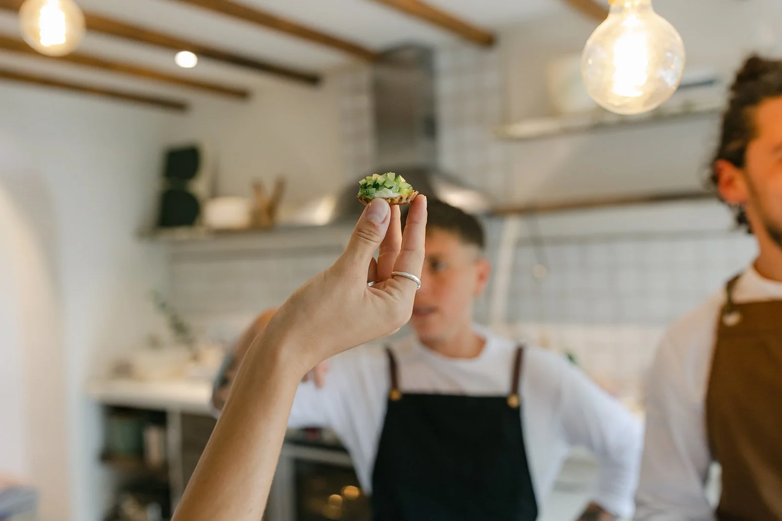 A person holding a small piece of food topped with chopped green vegetables in focus, with two people in chef aprons in the background in a kitchen.