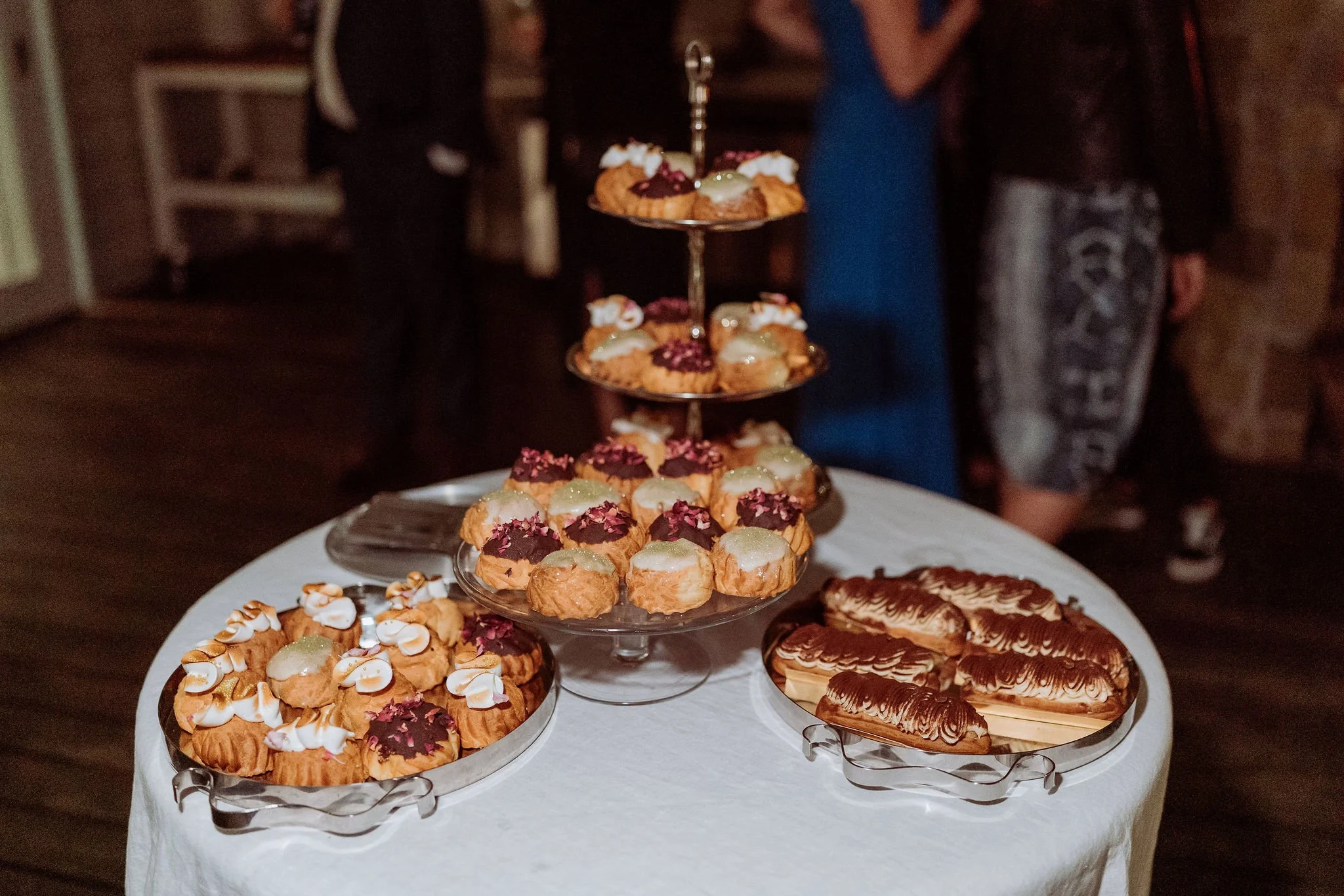 A table with a display of assorted small pastries and desserts, including cream puffs and eclairs, arranged on glass trays and a tiered stand, with a blurred background of people in a dimly lit room.
