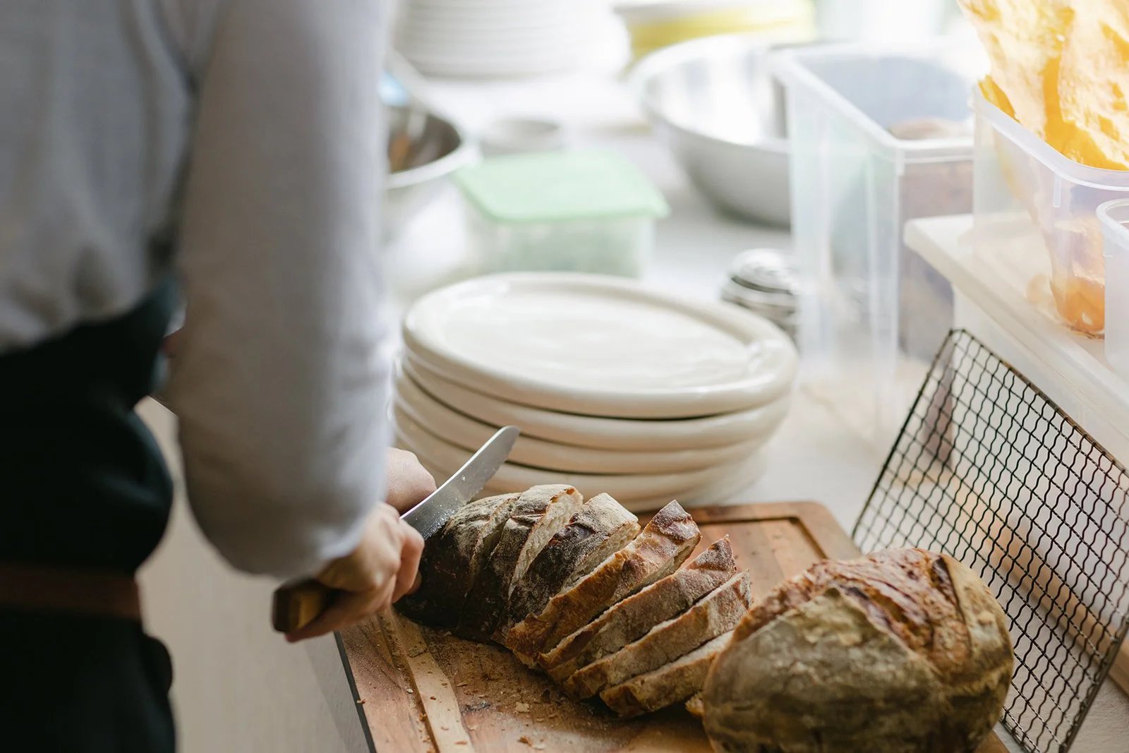 Person slicing a loaf of bread on a wooden cutting board in a kitchen with white plates and food containers in the background.