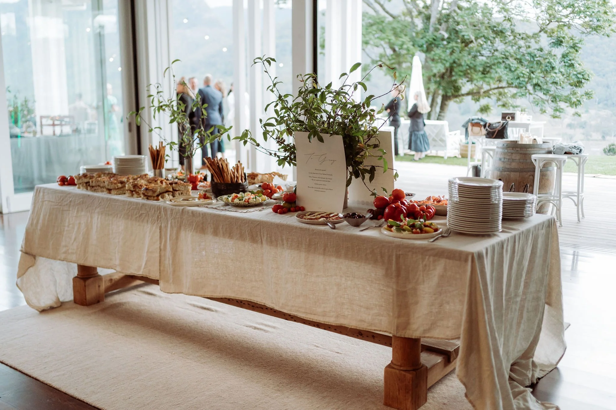 Buffet table with assorted food, fruits, and a plant centerpiece at a daytime outdoor event, with guests in the background.