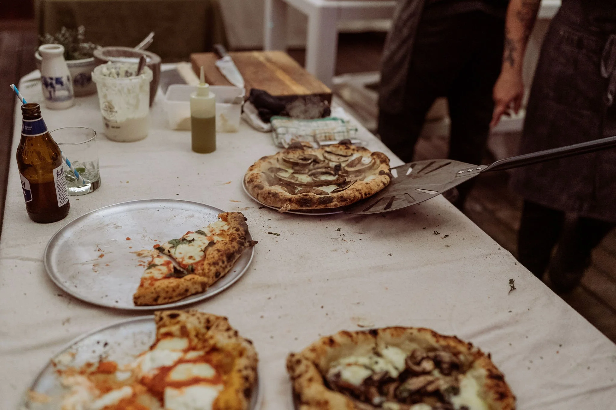 Leftover pizzas on plates and a pizza being served on a table covered with a white tablecloth, with condiments and drinks nearby.