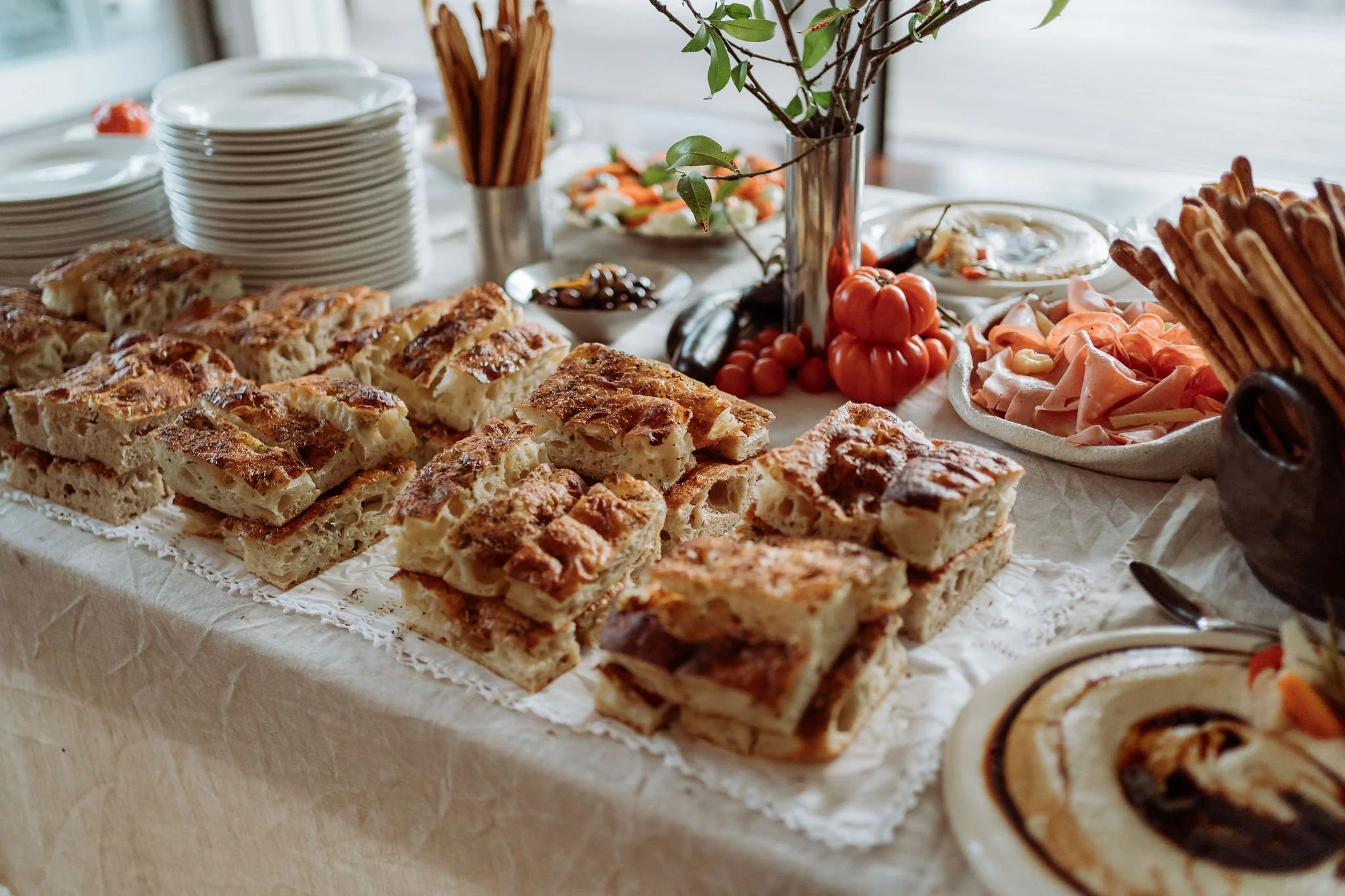 A table set with various baked goods including focaccia bread, a vegetable platter, a bowl of sliced meats, and a small pumpkin decoration, with plates and utensils in the background.