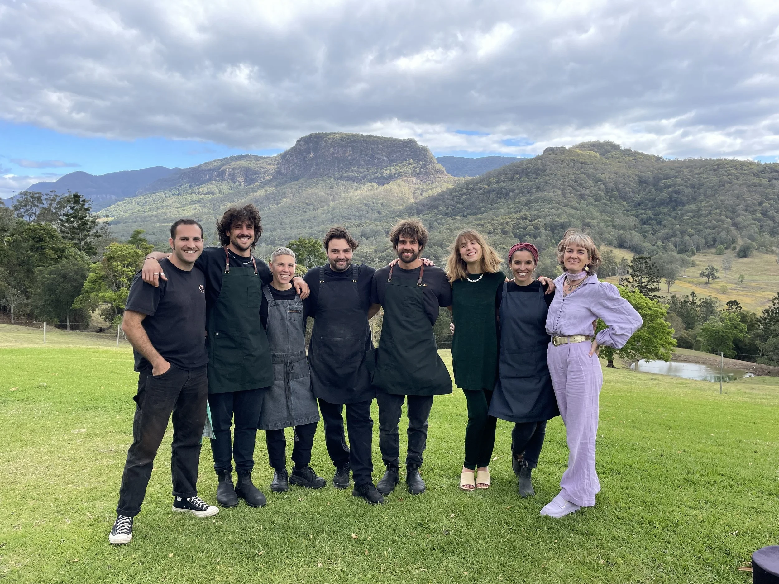 Group of seven people standing outdoors on green grass, smiling with their arms around each other, with mountains and trees in the background.