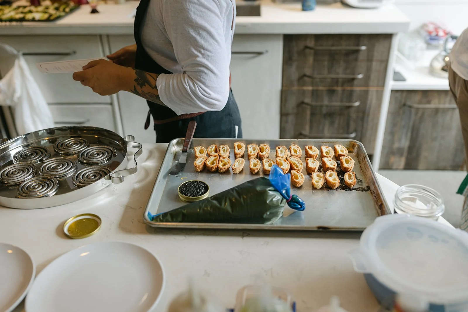 A person preparing sushi rolls on a tray with sliced pieces in a kitchen. There are ingredients and kitchen tools on the counter, including a jar of black caviar and a bag of seaweed.