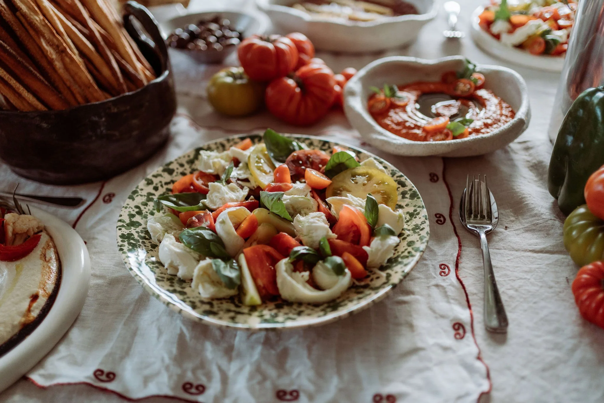 A rustic table spread with a plate of Caprese salad garnished with fresh basil, a bowl of tomato sauce, and various heirloom tomatoes, with a basket of breadsticks in the background.