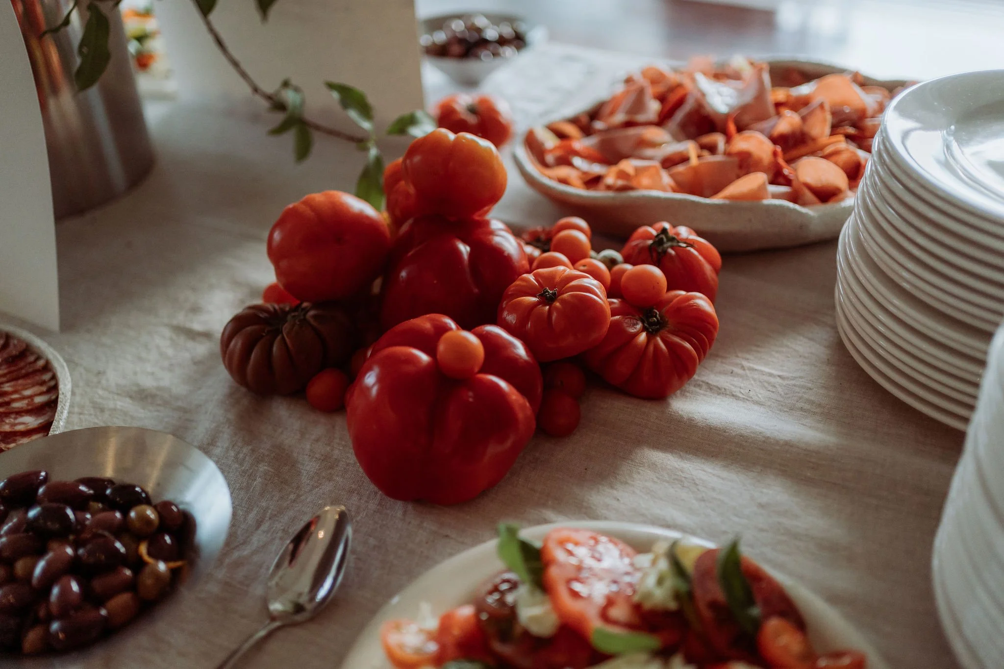 A table with heirloom tomatoes, a bowl of sliced peaches, a plate of pasta salad, and various plates and utensils.