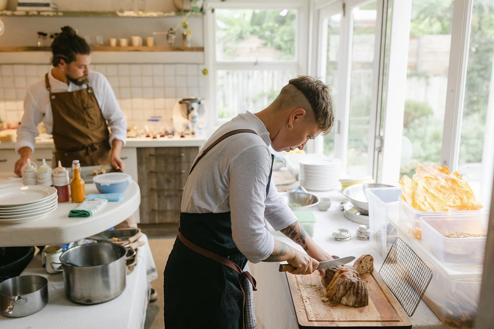 Two people in a bright kitchen preparing food; one is slicing a roast on a wooden cutting board, and the other is working at the counter in the background.