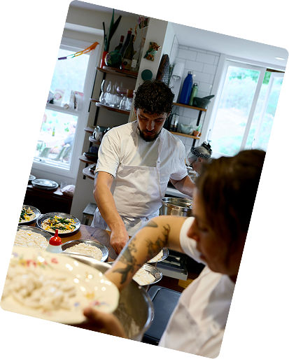 Two people cooking in a home kitchen, one with curly hair and the other with tattoos on their arm, preparing food.