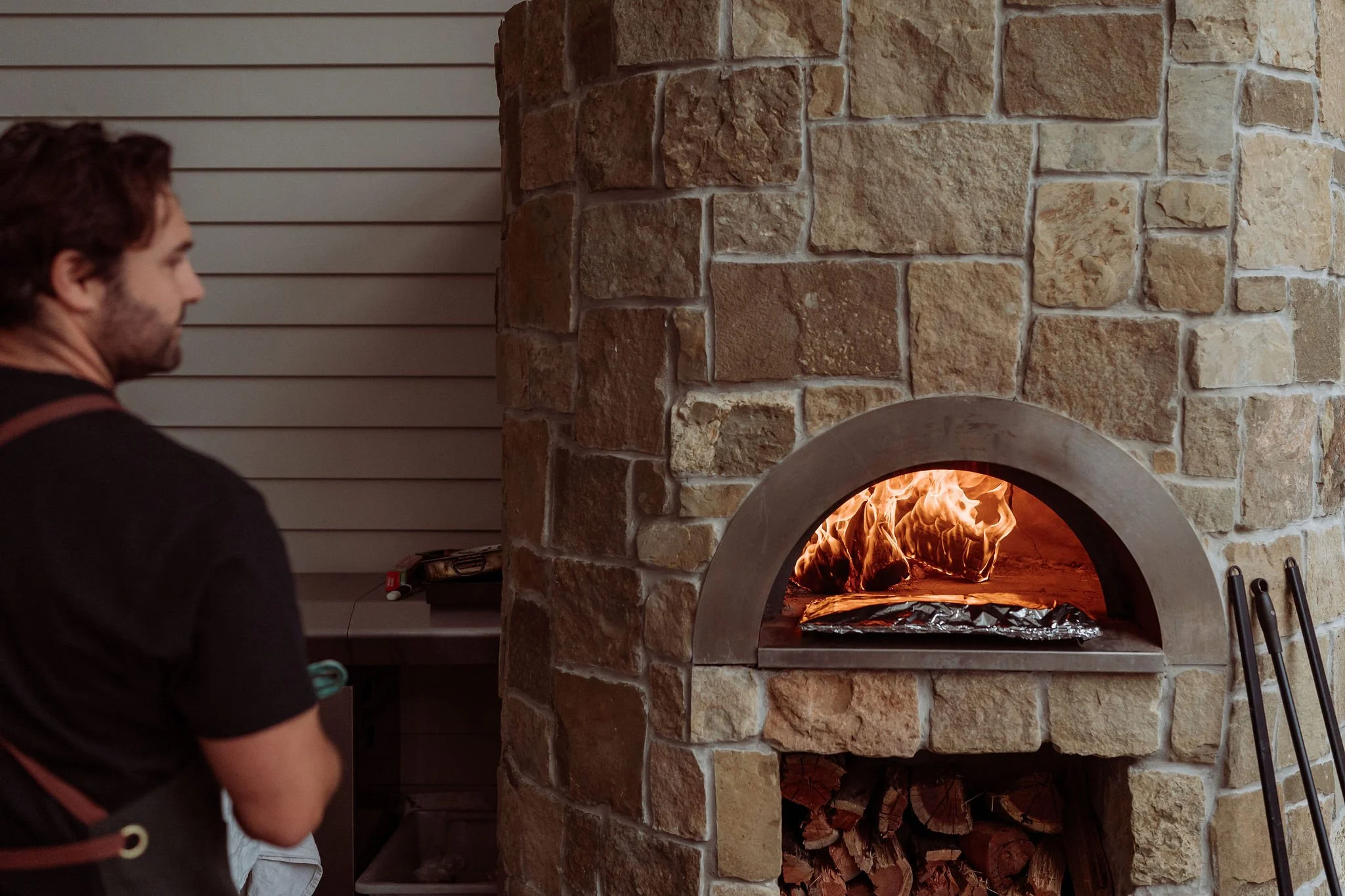 A man looks at a wood-fired pizza oven with visible flames inside, built with stone bricks, with firewood stored underneath.