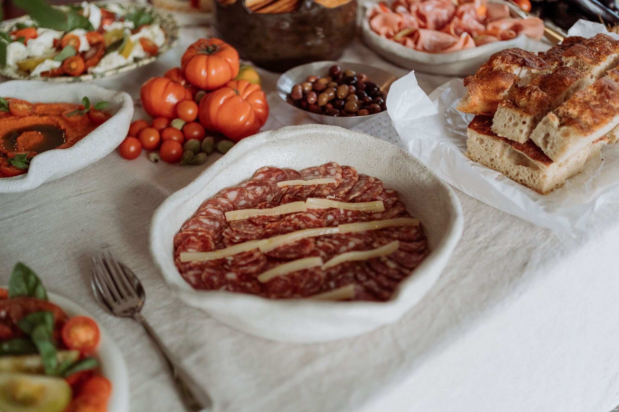 A variety of Italian antipasto dishes on a table, including sliced cured meat with cheese slices on top, fresh tomatoes, bread, and assorted vegetables.