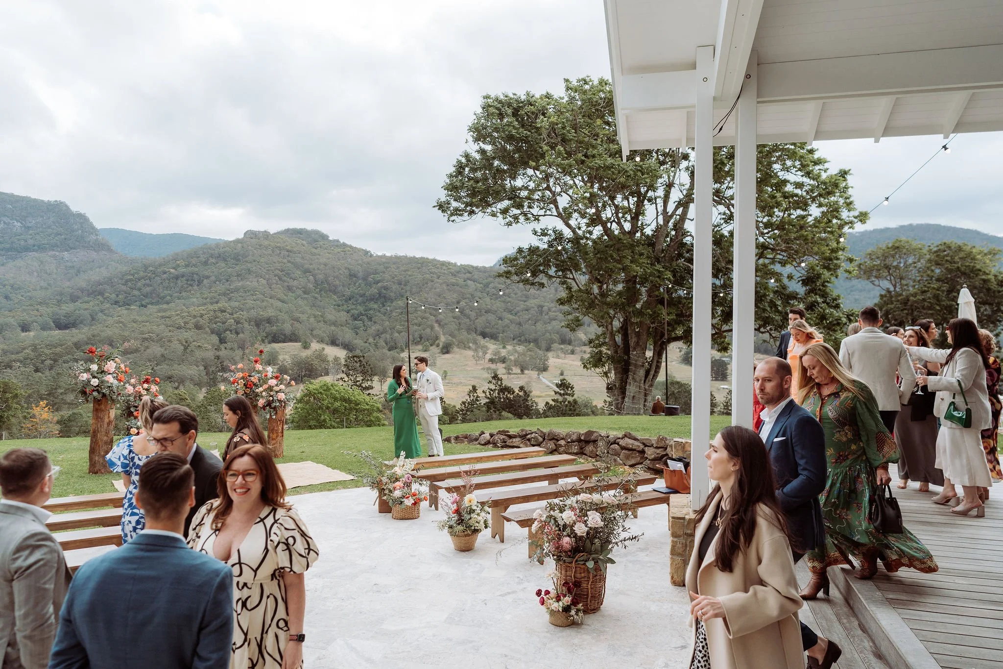A wedding ceremony taking place outdoors on a covered patio with guests mingling and a scenic mountain view in the background. The scene features floral decorations, including bouquets and arrangements, and a couple standing at the altar.