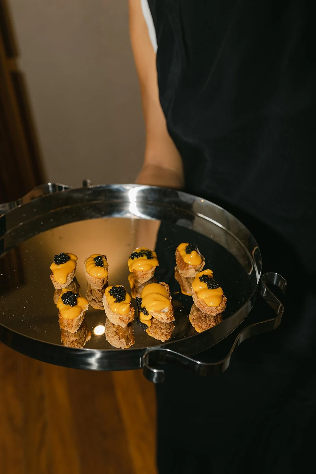 A person holding a silver tray with bite-sized appetizers topped with yellow sauce and black caviar.