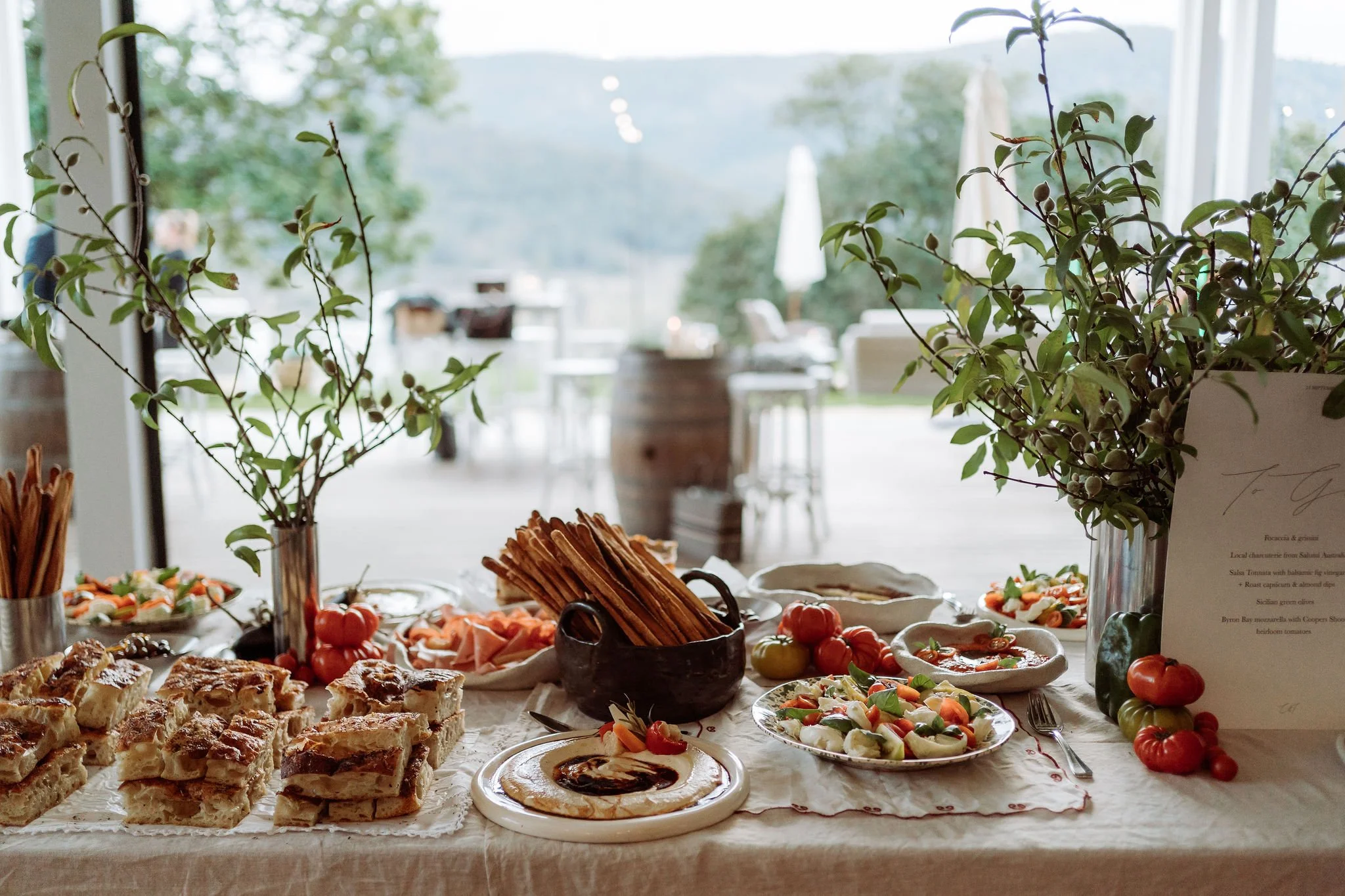 A table with food including pastries, salads, breadsticks, and fresh vegetables. Two vases with green leafy branches are on either side of the table, with a blurry outdoor patio in the background.