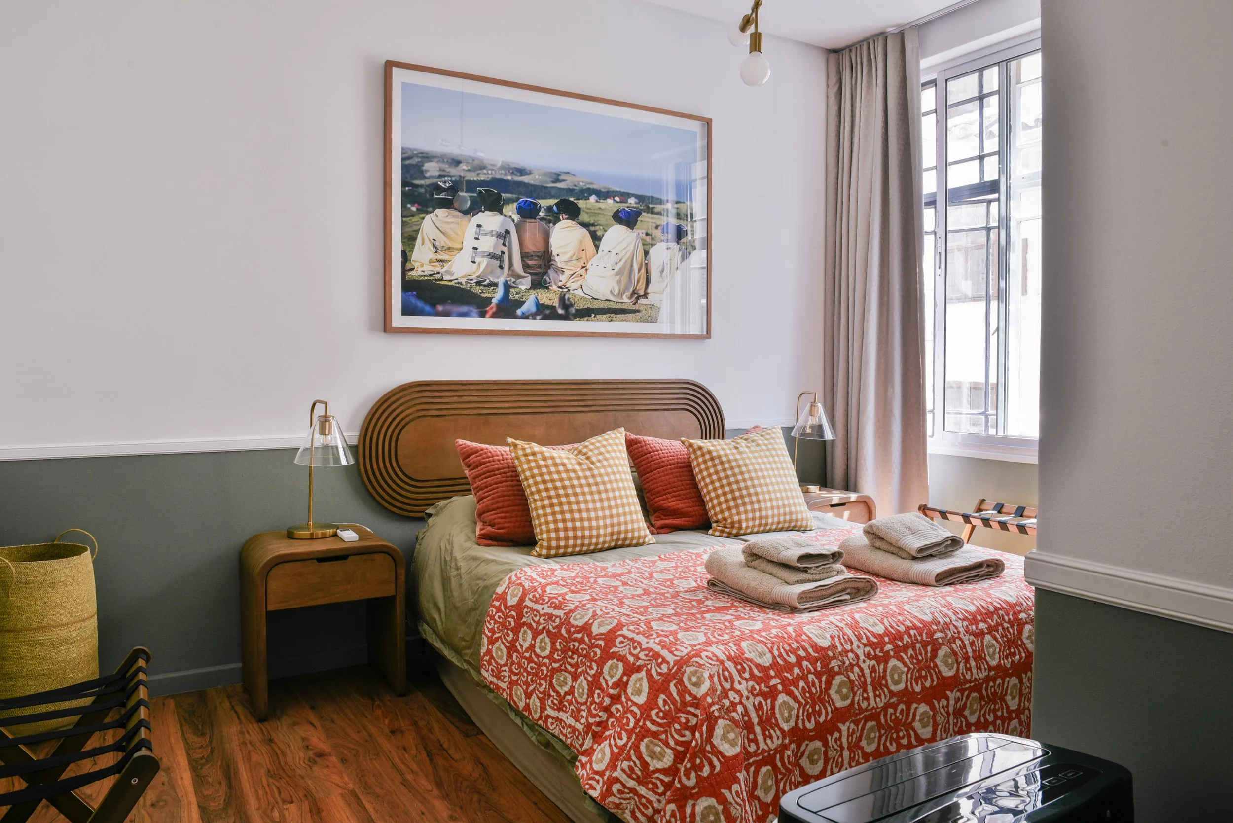 A neatly made bed with orange and yellow checkered pillows, a red patterned bedspread, and folded white towels on top. There are two bedside lamps on white nightstands, a wooden headboard, a large framed landscape photograph above, and a window with beige curtains allowing natural light into the room.