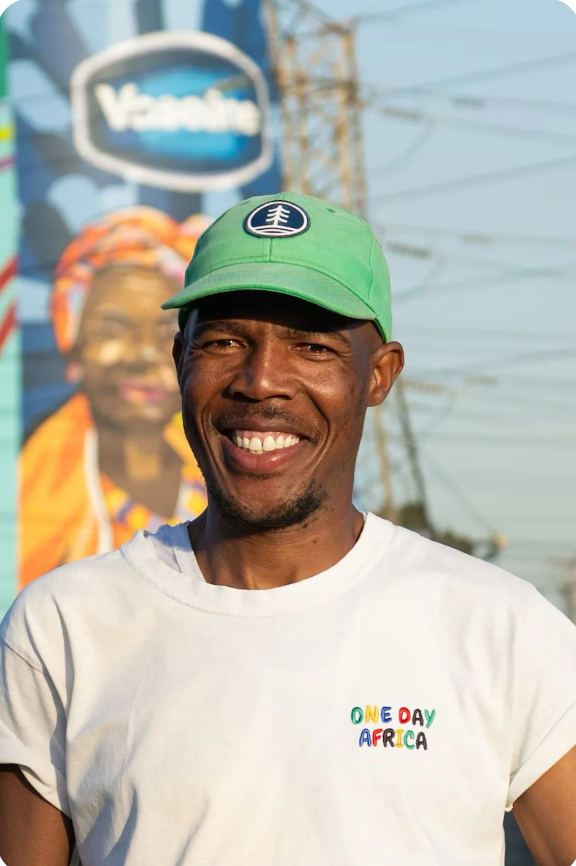 Smiling man wearing a green cap and a white t-shirt that says "One Day Africa" in colorful letters, with a background of African art and power lines.