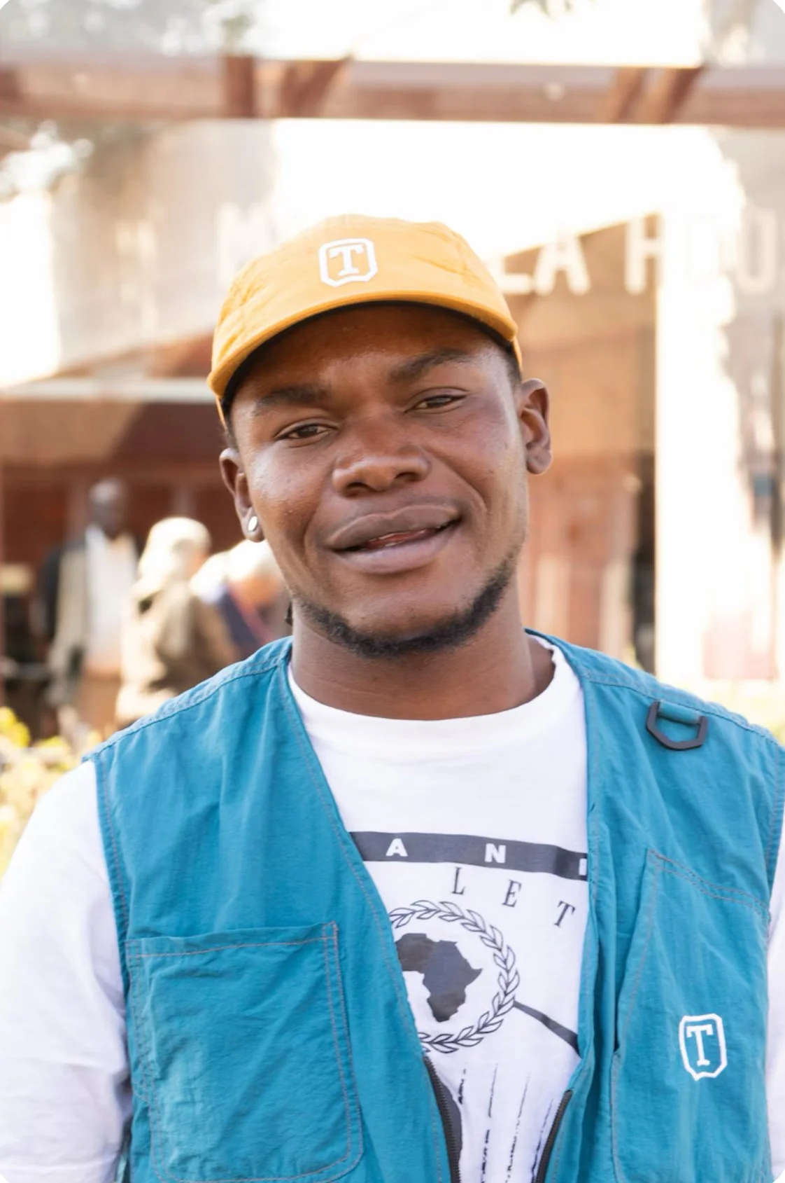 A young man wearing a yellow cap and a blue vest, standing outdoors with a blurred background.