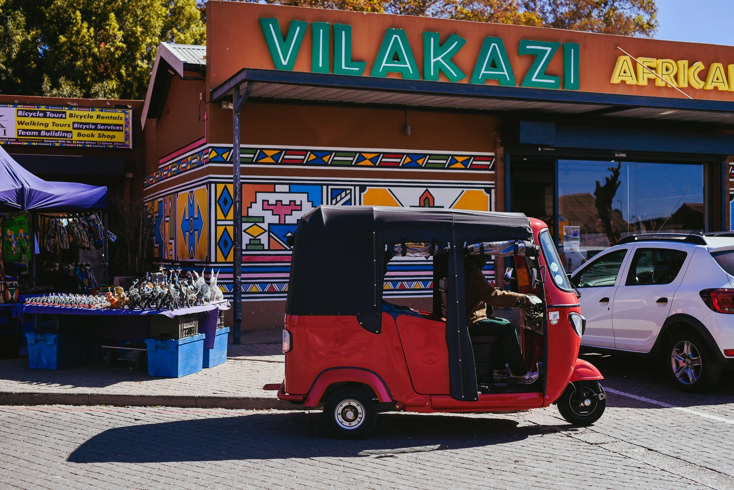 A red tuk-tuk parked on a street in front of a colorful African craft shop, Vilakazi African, with a large sign, beneath a mural with geometric patterns. A white car is nearby, and a vendor sells small animal figurines at a table covered with a purple cloth.
