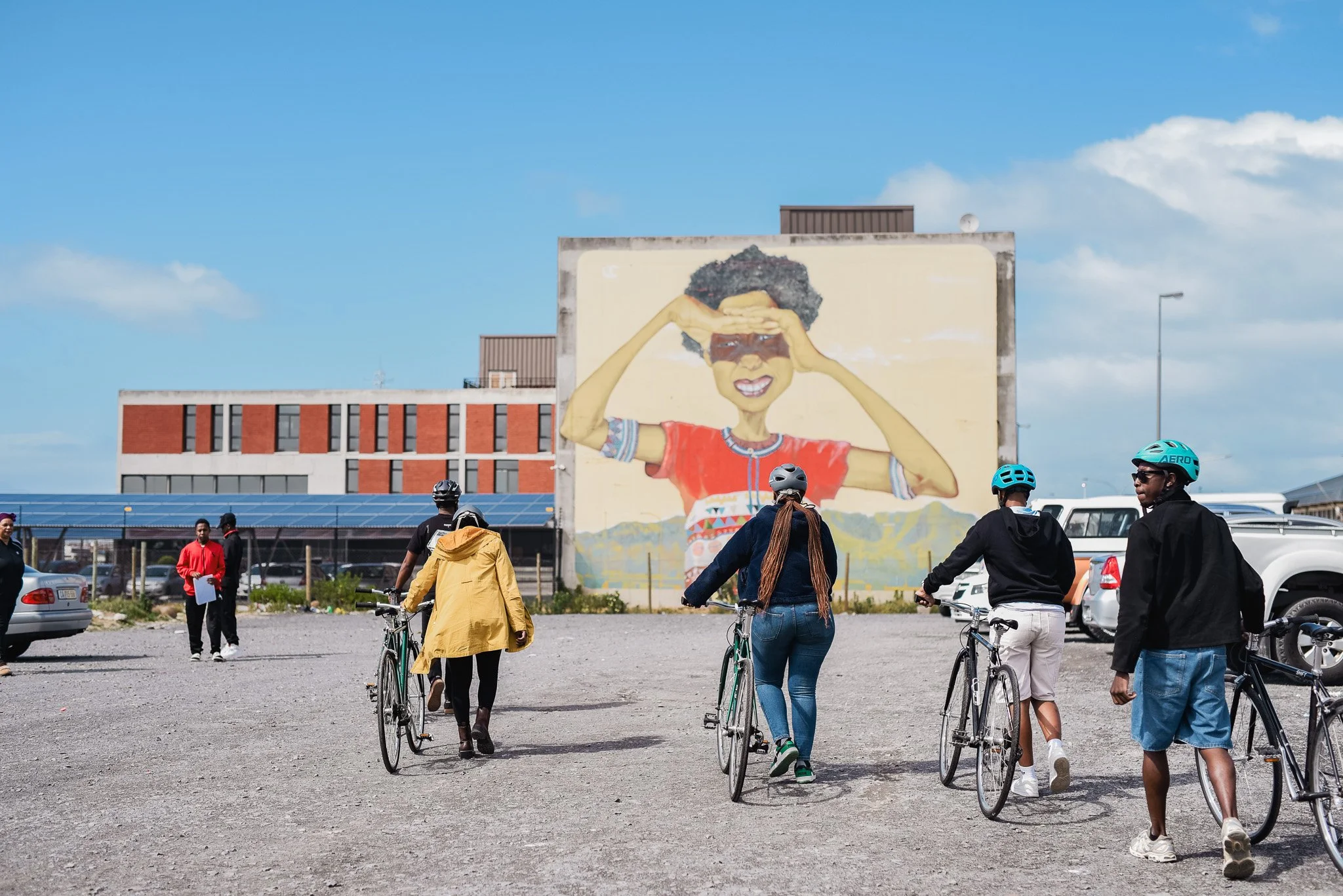 A group of five people riding bicycles towards a large mural of a woman with curly hair, wearing sunglasses, a red shirt, and smiling in an urban parking lot on a sunny day.