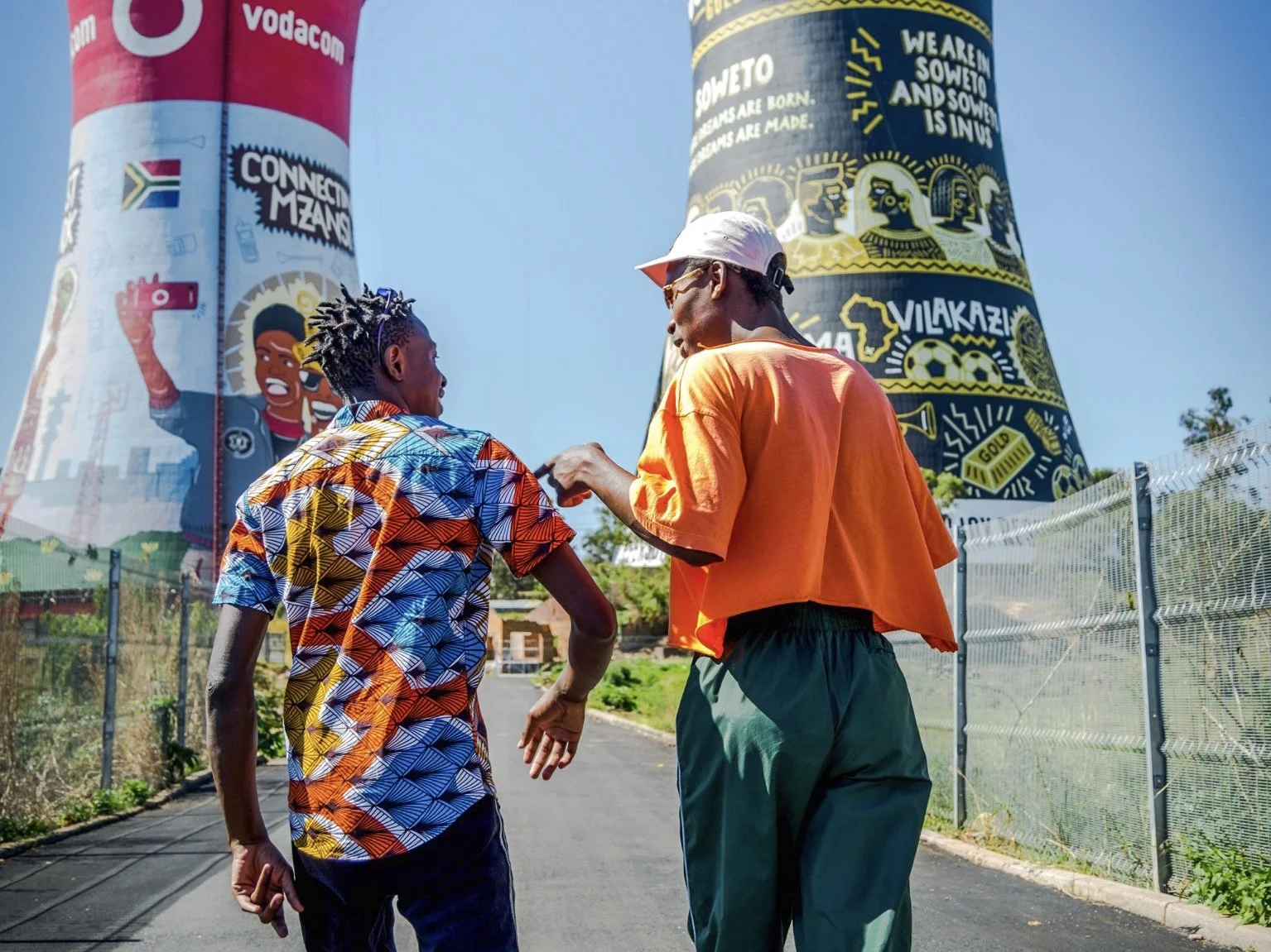 Two young men engaging in a fist bump outdoors near large colorful muraled towers against a clear blue sky.