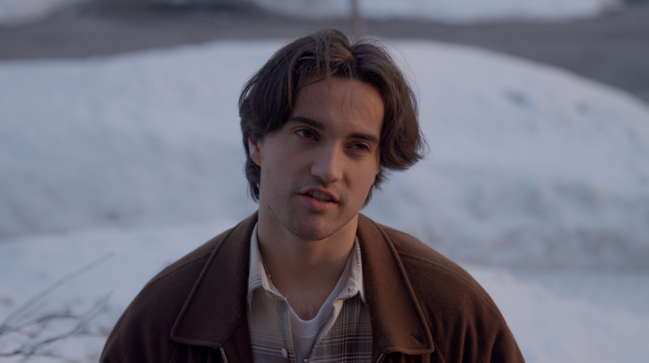 A young man with dark hair, wearing a brown jacket and plaid shirt, stands outdoors near snow-covered ground.