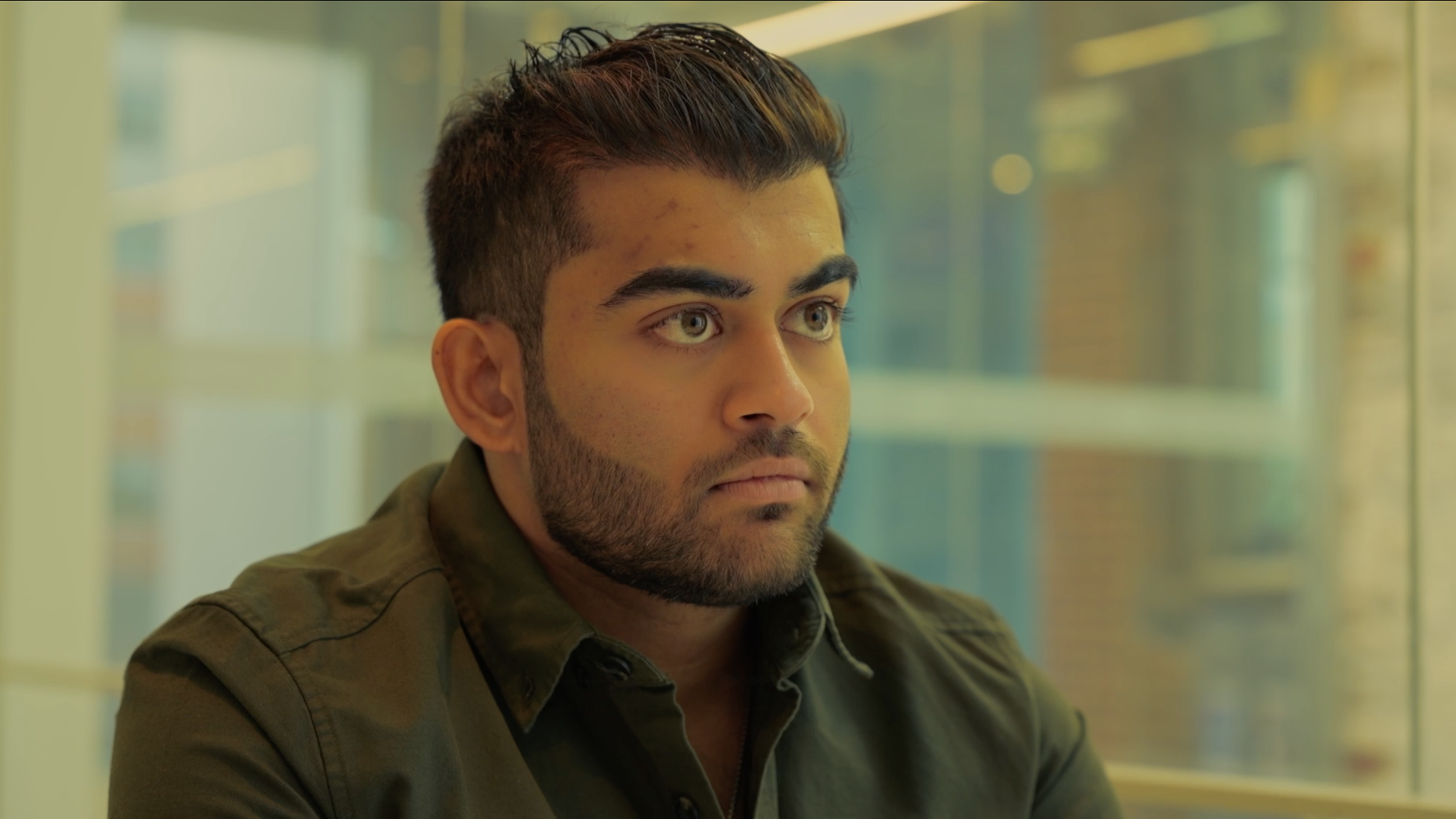 A young man with dark hair styled upward and gray-green eyes, wearing an olive green jacket, sitting indoors with a serious expression, blurred yellow and green background.