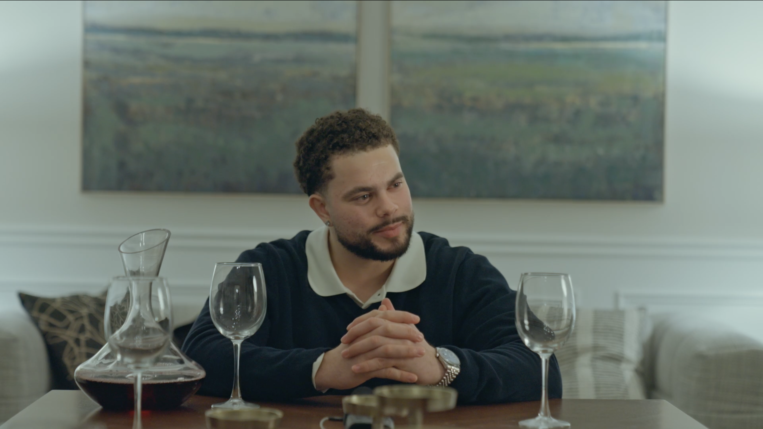 A young man with curly hair and a beard sits at a dining table with wine glasses and a decanter, looking thoughtfully to the side, with a large landscape painting in the background.
