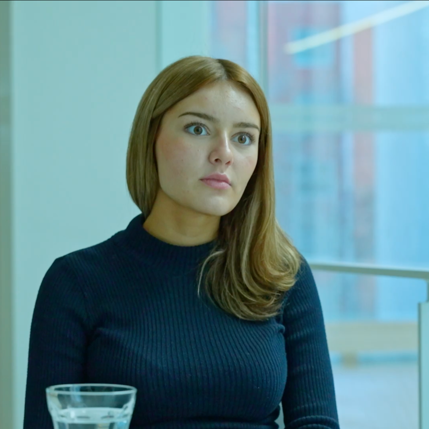 A young woman with long, styled brown hair and blue eyes, wearing a dark blue ribbed turtleneck sweater, sitting indoors near a large window with blurred cityscape outside.