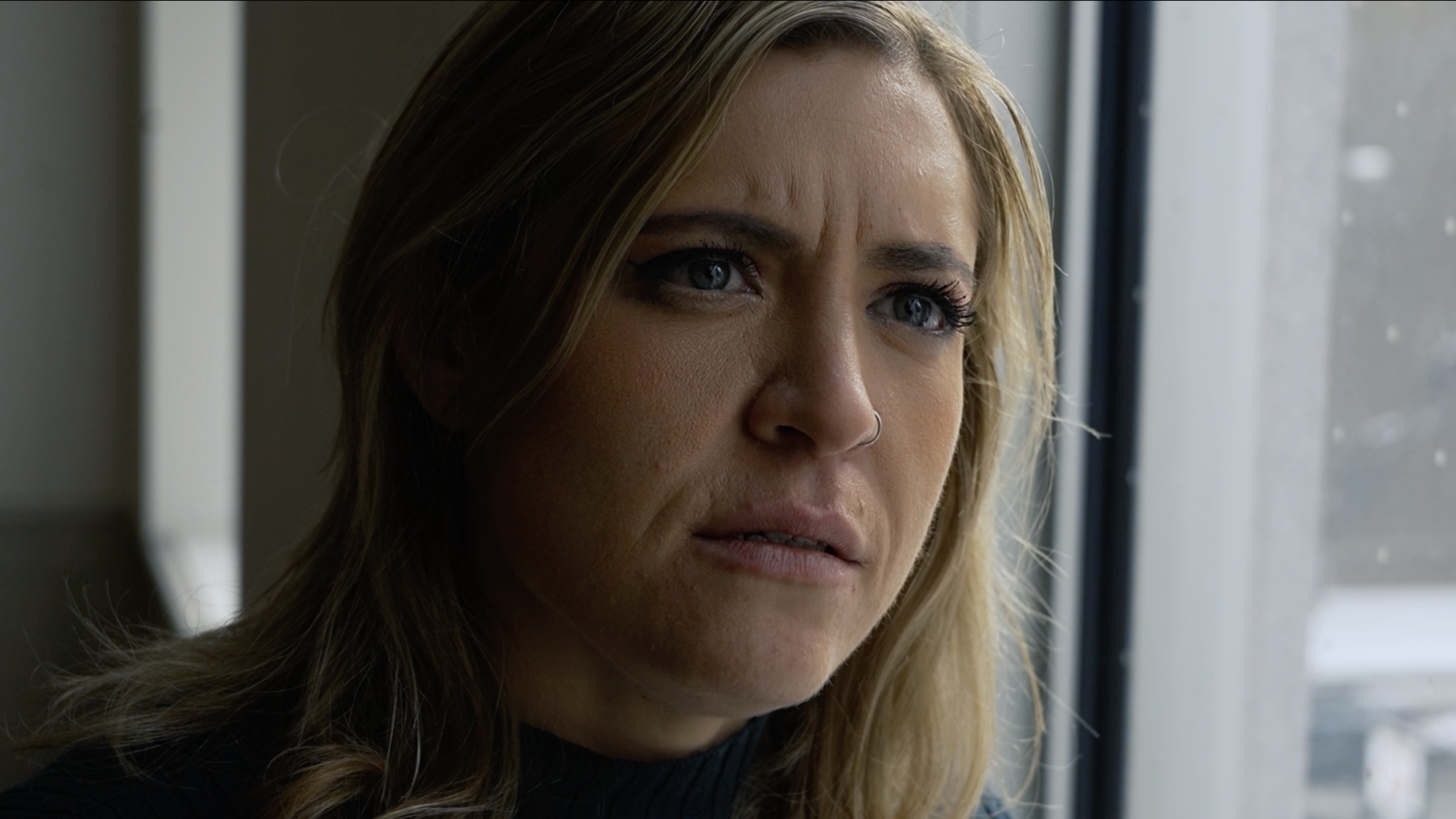 Close-up of a woman with blonde hair and blue eyes looking out a window.