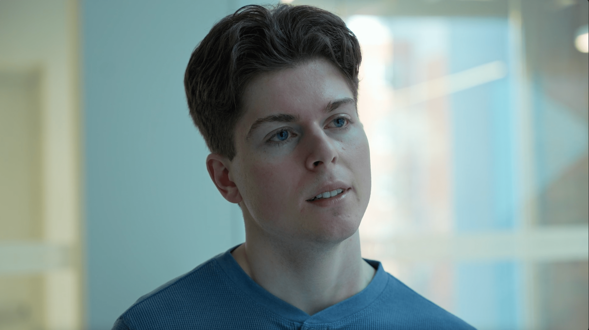 Close-up of a young man with short dark hair and blue eyes, wearing a blue top, in a brightly lit indoor setting.